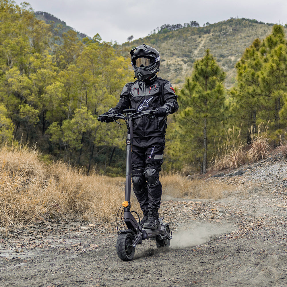 A person in full black riding gear and a helmet is riding a black electric scooter on a dirt path outdoors, with trees and hills in the background.