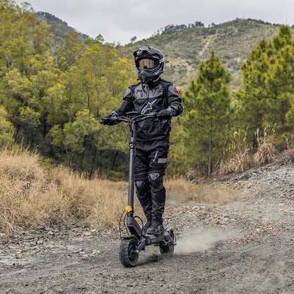 A person in full black riding gear and a helmet is riding a black electric scooter on a dirt path outdoors, with trees and hills in the background.