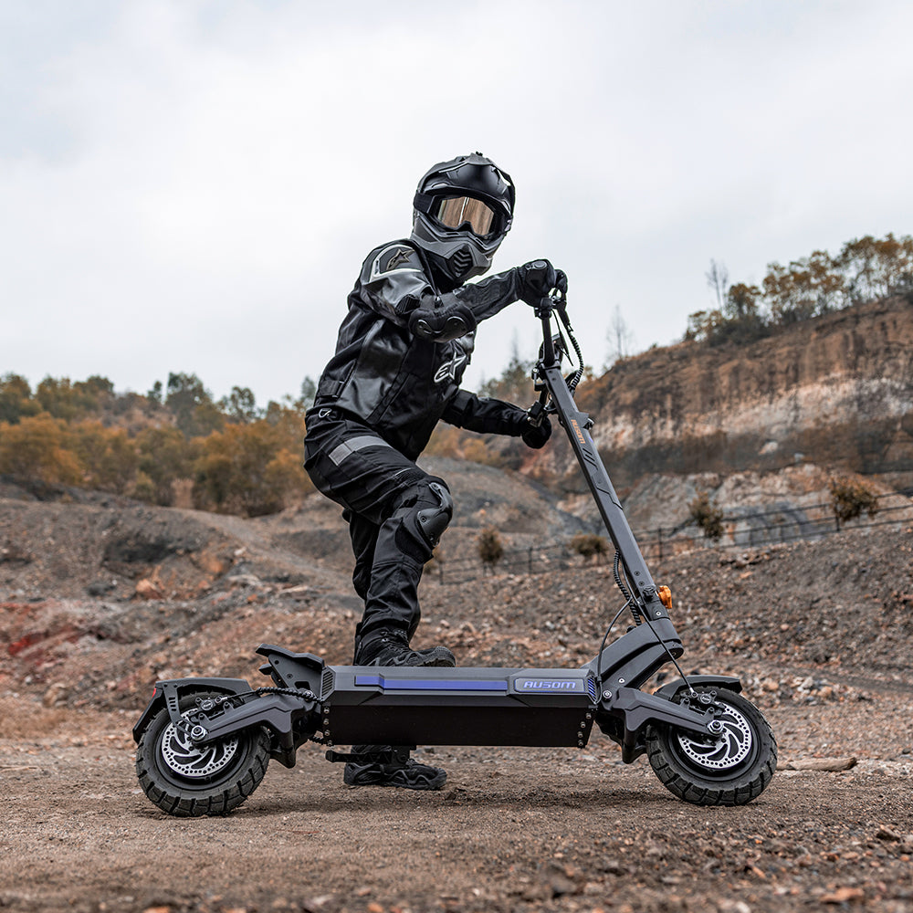 A person in full black riding gear and a helmet stands on a black electric scooter with knobby tires in an outdoor rocky, barren landscape under an overcast sky.