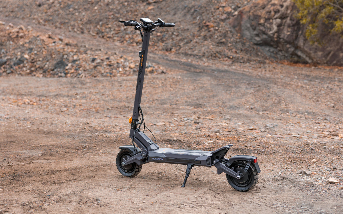 A dark gray electric scooter with a minimalist deck and upright stands parked on a dirt and gravel path outdoors.
